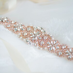 May include: A rose gold belt featuring an intricate arrangement of clear faceted crystals and small white pearls. The embellishment is set in a floral-inspired pattern on a rose gold base, creating a continuous decorative band of sparkling stones.