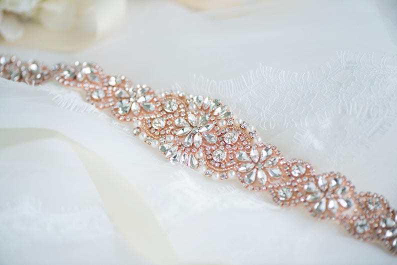 May include: A rose gold belt featuring an intricate arrangement of clear faceted crystals and small white pearls. The embellishment is set in a floral-inspired pattern on a rose gold base, creating a continuous decorative band of sparkling stones.