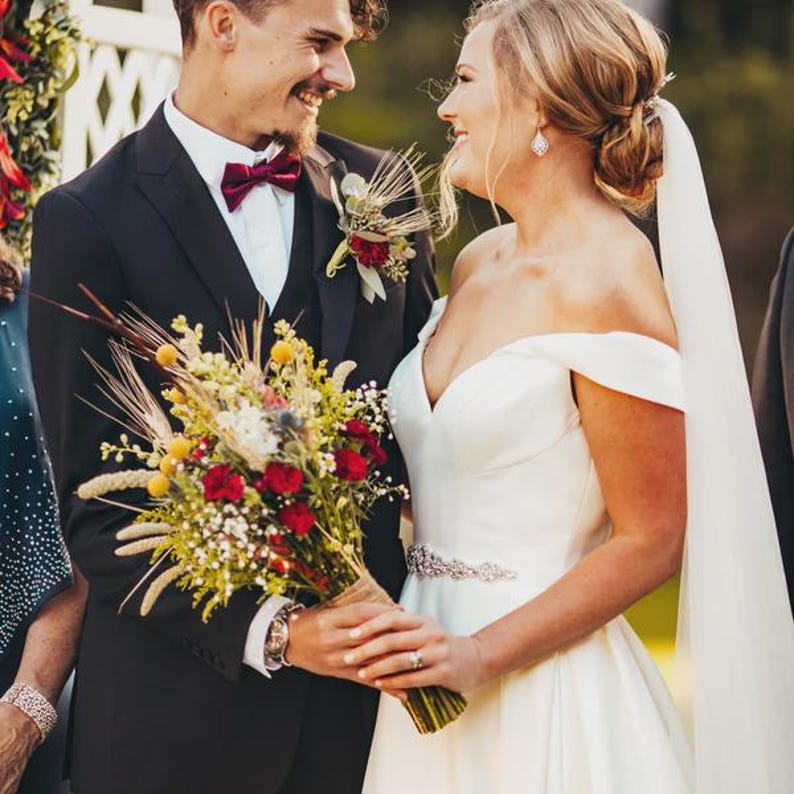 May include: A wedding couple in formal attire. The person on the left wears a black suit with a white shirt and a dark red bow tie. The person on the right wears an off-the-shoulder white gown with a beaded waist and a long veil.