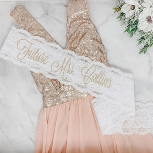 May include: A sleeveless dress with a rose gold sequined bodice and a flowing light pink skirt, draped with a white lace sash featuring the gold glitter text Future Mrs. Collins in a cursive script font, positioned diagonally across the bodice.