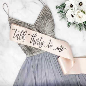 Puede incluir: Una faja de sat&eacute;n rosa claro con el texto "Talk thirty to me" escrito en caligraf&iacute;a negra. La faja est&aacute; atada alrededor de un vestido gris con lentejuelas.