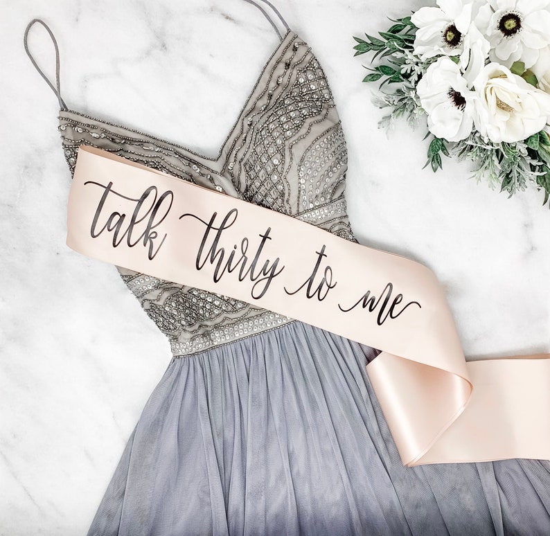 Puede incluir: Una faja de sat&eacute;n rosa claro con el texto "Talk thirty to me" escrito en caligraf&iacute;a negra. La faja est&aacute; atada alrededor de un vestido gris con lentejuelas.