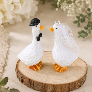 May include: Two white goose figurines standing on a circular wood slice. The left goose wears a black bowler hat and a black bow tie. The right goose wears a small silver crown and a white lace veil. Both geese have orange feet and beaks.