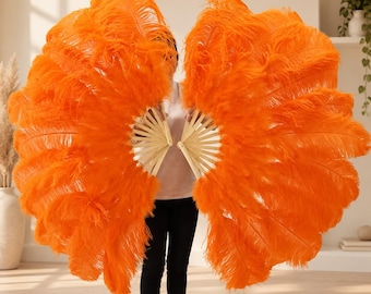 May include: A pair of large, vibrant orange ostrich feather fans featuring long, fluffy plumes attached to light-colored wooden ribs. The fans are held open in a wide, semi-circular shape, showcasing the dense, soft texture of the feathers.