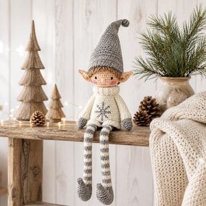 May include: A crocheted elf doll with a tall gray pointed hat, a cream-colored sweater featuring a gray snowflake design, and long gray and white striped legs. The doll has a light skin tone, pointed ears, and a small smile, sitting upright.