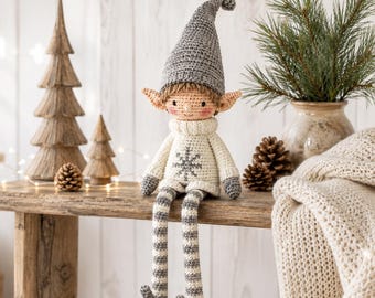 May include: A crocheted elf doll with a tall gray pointed hat, a cream-colored sweater featuring a gray snowflake design, and long gray and white striped legs. The doll has a light skin tone, pointed ears, and a small smile, sitting upright.