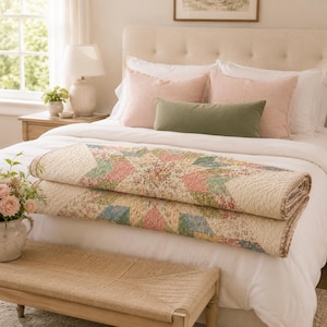 May include: A folded quilt featuring a multi-colored star pattern with floral prints in shades of pink, blue, green, and yellow on a cream background. The quilt is layered on a bed with white bedding, two pink pillows, and one olive green bolster.