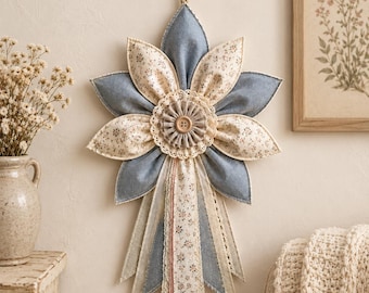 May include: A wall hanging featuring a fabric flower with alternating blue and floral-patterned petals, a central beige button, and a cream-colored lace doily. Below the flower, several hanging fabric ribbons in blue, floral, and lace textures.