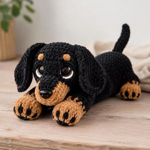 Op de afbeelding: Een gehaakt teckel knuffeltje gemaakt van zacht zwart en bruin garen. De hond heeft grote, glanzende zwarte ogen, een kleine zwarte neus, lange slappe oren en een lang lichaam met een omhoogstaande staart. De pootjes zijn bruin met zwarte details.