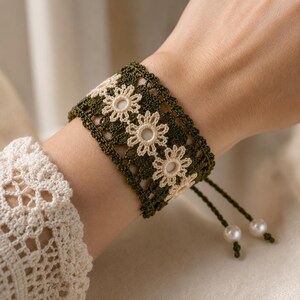 May include: A wide crochet bracelet featuring a dark olive green thread base with a central row of cream-colored flower motifs. The bracelet includes two adjustable drawstrings finished with small, round, white pearl-like beads at the ends.