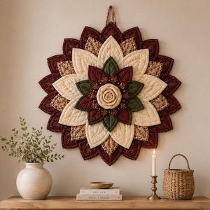 Puede incluir: Un tapiz circular de tela con un diseño de flor acolchada en capas en tonos burdeos intenso, crema y verde bosque. El centro es una rosa de tela enrollada, rodeada de pétalos concéntricos con costuras y patrones detallados.