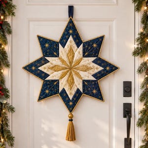 Op de afbeelding: Een gewatteerde wandhanger in de vorm van een achtpuntige ster in marineblauw, crème en goud met geborduurde stermotieven. De ster hangt aan een donkerblauw lint en is afgewerkt met een gouden kwastje aan de onderste punt.
