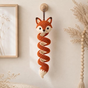 Peut inclure: Une décoration suspendue en crochet représentant un renard, avec une tête en fil orange et crème, attachée à une longue queue en spirale faite de mailles alternées orange et crème. Le renard est suspendu par un cordon marron à un crochet en bois.