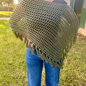 May include: An olive green crochet poncho featuring an open, grid-like stitch pattern and a wide, boat-style neckline. The garment is finished with long, matching olive green fringe tassels along the bottom edge, creating a textured, layered look.