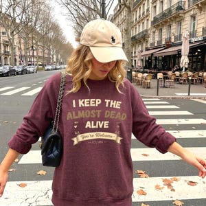 May include: A maroon crewneck sweatshirt featuring the text I KEEP THE ALMOST DEAD ALIVE You're Welcome - ICU RN - in cream and tan lettering. The sweatshirt is paired with a beige baseball cap with a small dark logo and a black quilted shoulder bag.