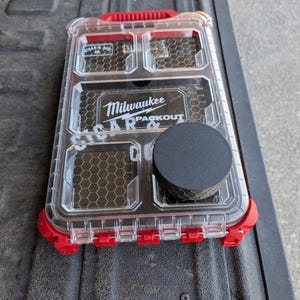 May include: A rectangular Milwaukee Packout storage organizer with a clear lid and red plastic frame. The lid features a honeycomb pattern, the Milwaukee logo, and the text Cigar & Ash Co. A solid black circular disc sits on the clear lid surface.