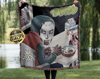 May include: A woven throw blanket featuring a graphic illustration of a masked figure in a hoodie eating cereal from a bowl labeled The Finest. The design includes a carton of cream, city buildings, and the text DOOM in a stylized green font.