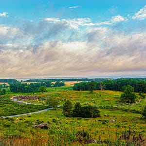 Op de afbeelding: Een panoramisch uitzicht op een grasveld met een kronkelende zandweg erdoorheen. De lucht is een levendig blauw met pluizige witte wolken. Het veld is bezaaid met bomen en struiken, wat een schilderachtig landschap creëert.