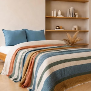 Könnte beinhalten: Ein Bett mit zwei einfarbigen blauen Kissen und einer strukturierten Tagesdecke mit vertikalen Streifen in Terrakotta, Senfgelb, Creme und gedämpftem Blau. Die Decke hat einen ausgefransten Rand und liegt auf der Matratze eines Holzbettrahmens.