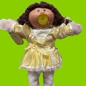 Könnte beinhalten: Eine Cabbage Patch Kids Puppe mit braunen Wollhaaren in Zöpfen, bekleidet mit einem gelben Satinkleid mit weißer Spitzenborte, einer gelben Schleife in der Taille, weiß gemusterten Strumpfhosen und weißen Schuhen. Ein gelber Schnuller im Mund.