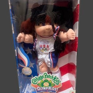 May include: A Cabbage Patch Kids Olympikids special edition doll in original packaging. The doll has dark yarn hair, purple eyes, and wears a white tank top with '1996 Olympikids USA' and Olympic rings, plus blue and white athletic shorts.