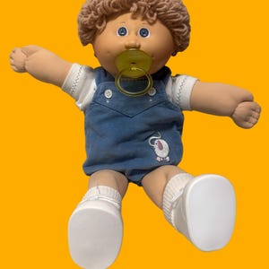 May include: A soft-bodied doll with light brown yarn hair and blue eyes, wearing a white short-sleeved shirt under blue corduroy overalls. The overalls feature a small embroidered elephant on the front. The doll has a yellow pacifier and white shoes.