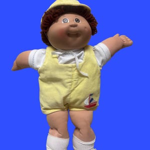 Op de afbeelding: Een pop met bruin garen haar, een gele pet, een wit shirt met korte mouwen en een gele ribfluwelen tuinbroek met een klein geborduurd zeilbootje. De pop draagt witte sokken en witte veterschoenen en heeft een rond, zacht gevormd gezicht.