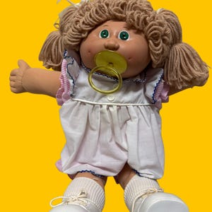 May include: A doll with light brown yarn hair styled in pigtails, green eyes, and a tan complexion. The doll wears a white dress with pink striped ruffled sleeves, a yellow pacifier in its mouth, white ribbed socks, and white shoes.