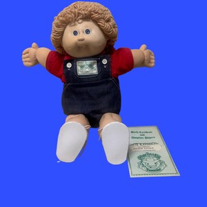 Può includere: Una bambola Cabbage Patch Kids con capelli in lana marrone chiaro e occhi blu, che indossa una maglietta rossa sotto una salopette in denim blu scuro con bottoni bianchi. La bambola è accompagnata da un certificato di nascita e adozione.