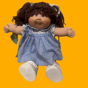 Könnte beinhalten: Eine Cabbage Patch Kids Puppe mit dunkelbraunen Wollhaaren in Zöpfen, blauen Augen und einem Gesicht mit Grübchen. Die Puppe trägt ein blau-weiß kariertes Kleid mit weißem Kragen und weißen Schuhen. Ein grünes Etikett ist am Arm befestigt.