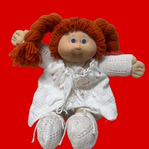 Puede incluir: Una muñeca de tela con cabello de lana naranja brillante peinado en dos coletas y ojos azules. La muñeca viste un cárdigan de punto blanco con pequeños detalles rosas, un vestido blanco estampado y patucos de punto blancos a juego.