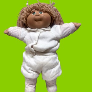 Puede incluir: Una muñeca con cara redonda, piel bronceada y ojos verdes, con cabello de lana marrón claro adornado con pequeñas cintas blancas. La muñeca viste una camisa blanca de manga larga con cuello y pantalones cortos blancos a juego, con mallas rosa claro y zapatos blancos.