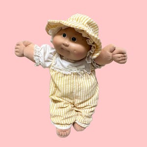 Puede incluir: Una muñeca de tela con cara redonda, hoyuelos y ojos oscuros, que viste una camisa blanca de manga corta con ribete de encaje, un peto a rayas amarillas y blancas y un sombrero de sol a juego a rayas amarillas y blancas con encaje.