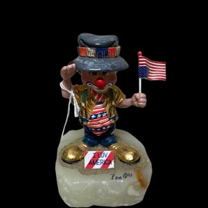 May include: A clown figurine wearing a grey hat, gold shirt, and blue pants, saluting while holding a small American flag. The clown stands on a stone base with a sign that reads I LUV AMERICA and a signature that says Ron 91 in black ink.