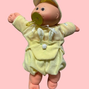 May include: A doll with a round face, peach-coloured skin, and dark eyes, wearing a pale yellow long-sleeved outfit with a front pocket featuring white rabbit ears and a small white pom-pom. The doll wears a matching yellow hat and a yellow pacifier.