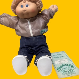 Può includere: Una bambola Cabbage Patch Kids con capelli in lana marrone chiaro, occhi blu e guance rosate. La bambola indossa una giacca beige con cerniera e logo verde, pantaloni in denim blu scuro e scarpe bianche. Accanto c'è un certificato di nascita.