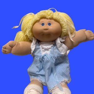 May include: A doll with yarn blonde hair in pigtails, blue eyes, and a tan complexion. The doll wears a light blue dress with white polka dots, white lace trim, and a beaded necklace with the letters spelling CHERYS around the neck.