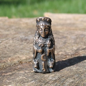 May include: A small, metallic bronze-toned figurine depicting a figure with a crescent moon crown, holding a key and a torch. The figure is adorned with a central crescent moon symbol and a key, flanked by two small animal figures at the base.