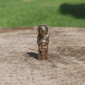 May include: A small, metallic, bronze-colored figurine depicting a standing figure with a stylized headdress. The figure has hands clasped over the chest and features a prominent circular emblem with a cross design centered on the lower torso.