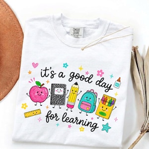 May include: A white t-shirt featuring a colorful graphic with the text 'it's a good day for learning'. The design includes cartoon illustrations of a pink apple, a black notebook, a yellow pencil, a blue backpack, a crayon box, and a ruler.