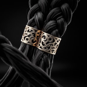 Peut inclure: Une barrette à cheveux en métal doré ornée d'un motif complexe de volutes en filigrane. L'accessoire est fixé autour d'une épaisse tresse de cheveux noirs, présentant un design ajouré détaillé avec de petites textures pointillées.