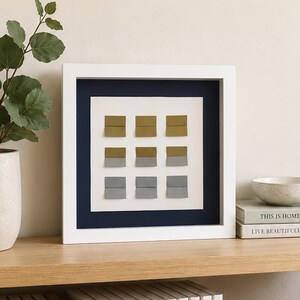 May include: A square wall art piece in a white frame featuring a navy blue mat and a white background. Nine small, folded rectangular elements are arranged in a three-by-three grid, transitioning in color from mustard yellow to light gray.