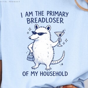 May include: A light blue t-shirt featuring a dark blue graphic of an opossum wearing sunglasses, holding a martini glass and a handbag. The text reads I AM THE PRIMARY BREADLOSER OF MY HOUSEHOLD. The design includes small star accents.