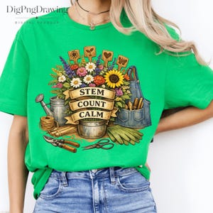 Pode incluir: Uma t-shirt verde brilhante com uma ilustração gráfica de ferramentas de jardinagem, um regador de metal, um avental azul e um ramo de flores coloridas. Uma faixa bege central exibe o texto STEM COUNT CALM em letras pretas.