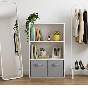 May include: A white three-tier bookshelf featuring two open shelves filled with books, a small potted plant, a reed diffuser, and a picture frame, plus two grey fabric storage bins at the base. The unit is positioned between a floor mirror and a clothing rack.
