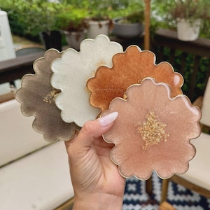 May include: A set of four flower-shaped coasters made of resin in shades of brown, cream, copper, and light pink. Each coaster features a smooth, scalloped edge and a central cluster of gold leaf flakes embedded within the glossy, semi-translucent material.