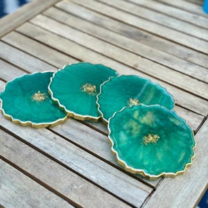 May include: Four circular coasters made of emerald green resin with a swirling, translucent finish. Each coaster features a textured gold-colored metallic edge and a central cluster of embedded gold leaf flakes, creating a geode-inspired appearance.