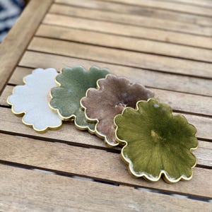 May include: A set of four flower-shaped coasters with a glossy finish and metallic gold edges. The coasters feature a smooth, swirling resin texture in shades of white, sage green, dark brown, and olive green, arranged in a slightly overlapping row.