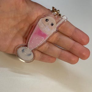 Puede incluir: Un llavero de acrílico transparente con forma de vaso alto lleno de una bebida rosa, cubierto con una bola de helado de color tostado claro con dos ojos oscuros. El diseño incluye una pajita blanca y una anilla de metal dorado en la parte superior.