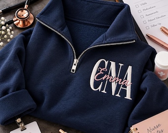 以下が含まれることがあります： 胸元に白い刺繍の文字「CNA」とピンクの筆記体で「Emma」と縫い込まれた、ネイビーブルーのクォータージッププルオーバー。ハイネック、袖口のある長袖、シルバーカラーの金属製ジッパーが特徴の衣類です。